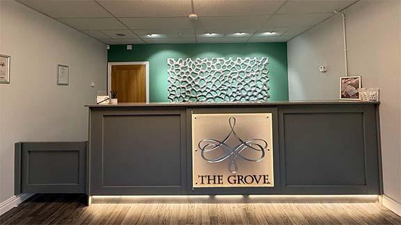 Reception desk in the evening, ceiling spotlights on, throwing light onto the Reception wall art, a large silver rectangle containing irregular molten bubble shapes. The centre desk panel has a backlit square sign with our chandelier logo on