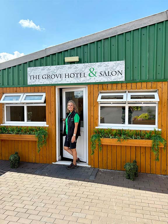 Lindsay at the Dog grooming salon entrance. There is a sign above the door saying 'The Grove Hotel and Salon'. There are plant troughs under the windows and it is a bright, sunny day